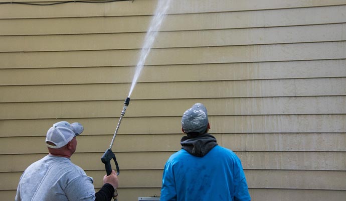 two young man standing by the side of a yellow house one is power washing the siding